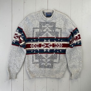 Könnte beinhalten: Ein grauer Strickpullover mit einem geometrischen Muster in Grautönen, Rot, Grün, Blau und Weiß. Der Pullover hat einen Rundhalsausschnitt und lange Ärmel.