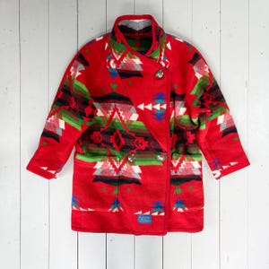 May include: A red wool coat with a Southwestern-inspired geometric pattern in shades of green, white, black, and blue. The coat features a double-breasted closure with two silver buttons, two front pockets, and a notched collar. A small blue tag is visible.