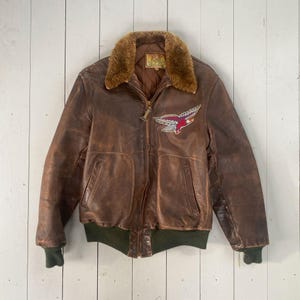 Puede incluir: Una chaqueta bomber de cuero marrón con cuello de piel y un bajo de punto de canalé verde. La chaqueta tiene una cremallera y un parche en el lado izquierdo con un diseño de águila bordado.