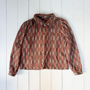 Può includere: Una camicia abbottonata con un motivo geometrico di rettangoli marroni, rossi e beige. La camicia a maniche lunghe ha un colletto e si allaccia con quattro bottoni rotondi color oro. La camicia è esposta su una superficie di legno bianco.