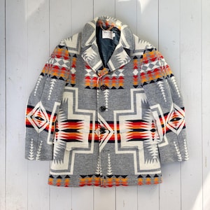 Op de afbeelding: Een grijze wollen jas met een geometrisch patroon in wit, rood, oranje en zwart. De jas heeft een reverskraag, drie knopen en twee voorzakken. De jas is van gemiddelde lengte.