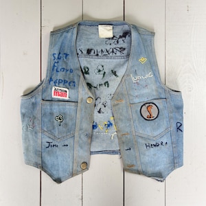 May include: A vintage denim vest with hand-painted details and patches. The vest features the words "Sgt. Floyd Pepper" and "Action Man" on the front, along with a shamrock and a cobra patch. Additional text includes "Jimi," "Hendra," and "Ludwig."