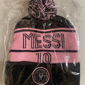 May include: Black and pink knit beanie with a pom pom on top. The beanie has the text "MESSI 10" on the front and a logo with the text "Club Internacional de Futbol Miami MMXX" on the back.
