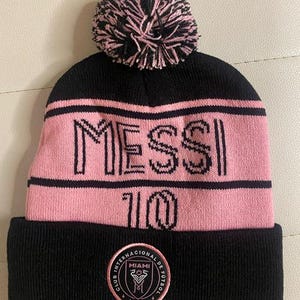 May include: Black and pink knit beanie with a pom pom on top. The beanie has the word "MESSI" and the number "10" on the front. The beanie also has a black patch with the words "Club Internacional de Futbol Miami" and the year "MMXX" on the back.