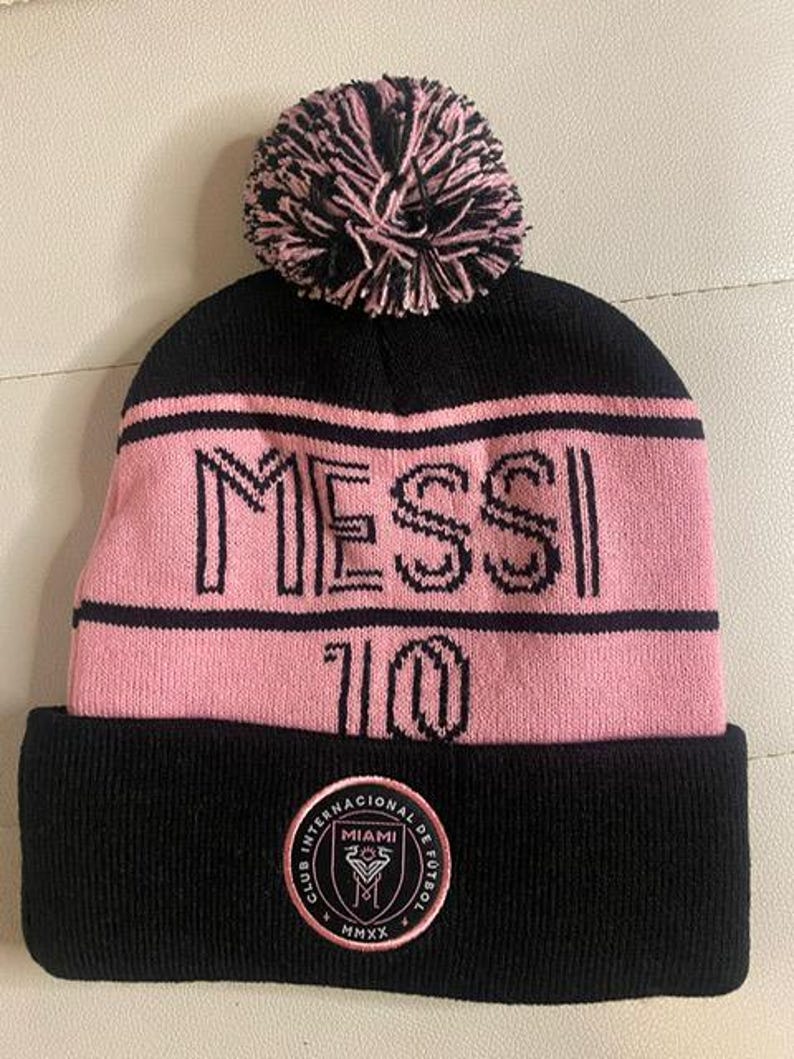 May include: Black and pink knit beanie with a pom pom on top. The beanie has the word "MESSI" and the number "10" on the front. The beanie also has a black patch with the words "Club Internacional de Futbol Miami" and the year "MMXX" on the back.