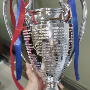 Puede incluir: Un trofeo plateado brillante con dos grandes asas y un cuenco ancho. El trofeo tiene una lista de nombres grabados en su lateral. Cintas rojas y azules est&aacute;n atadas a las asas.
