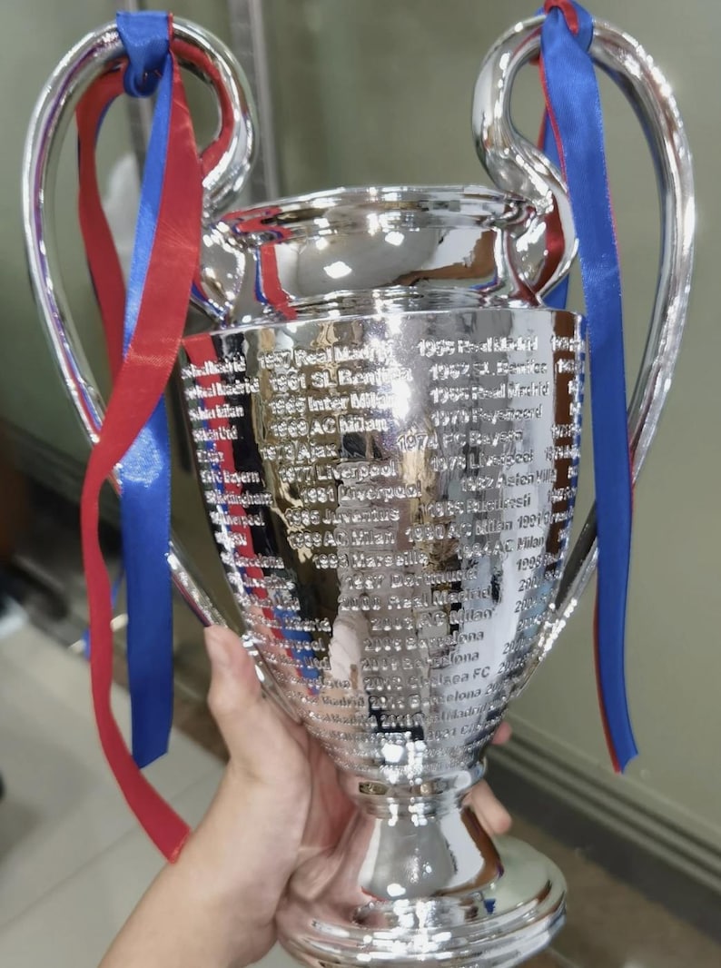Puede incluir: Un trofeo plateado brillante con dos grandes asas y un cuenco ancho. El trofeo tiene una lista de nombres grabados en su lateral. Cintas rojas y azules est&aacute;n atadas a las asas.
