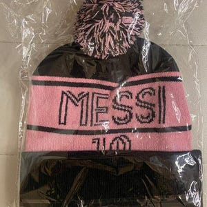 May include: Black and pink knit beanie with a fluffy pom pom and the word "MESSI" in black lettering.