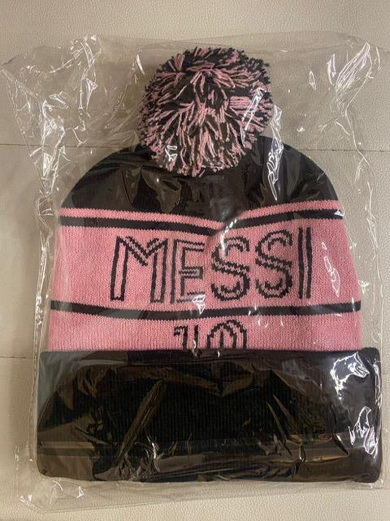 May include: Black and pink knit beanie with a fluffy pom pom and the word "MESSI" in black lettering.