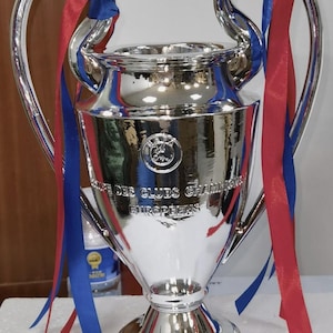 Puede incluir: Un trofeo plateado brillante con grandes asas y una base ancha. Cintas rojas y azules est&aacute;n atadas a las asas. El trofeo tiene grabadas las palabras "Des Clubs Champions Europeens".