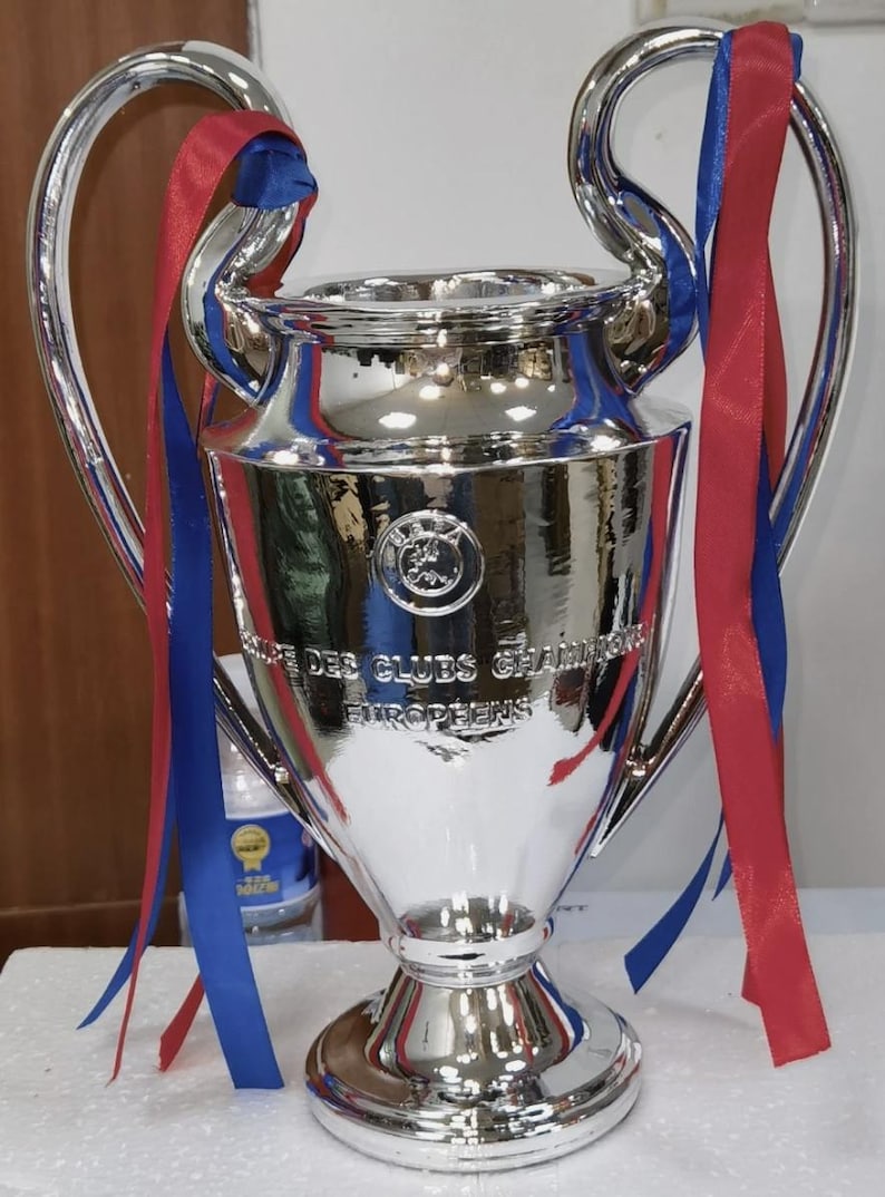 Puede incluir: Un trofeo plateado brillante con grandes asas y una base ancha. Cintas rojas y azules est&aacute;n atadas a las asas. El trofeo tiene grabadas las palabras "Des Clubs Champions Europeens".
