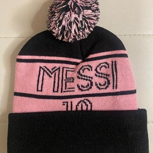 May include: Black and pink knit beanie with a pom pom on top. The beanie has the word "MESSI" and the number "10" embroidered on the front.
