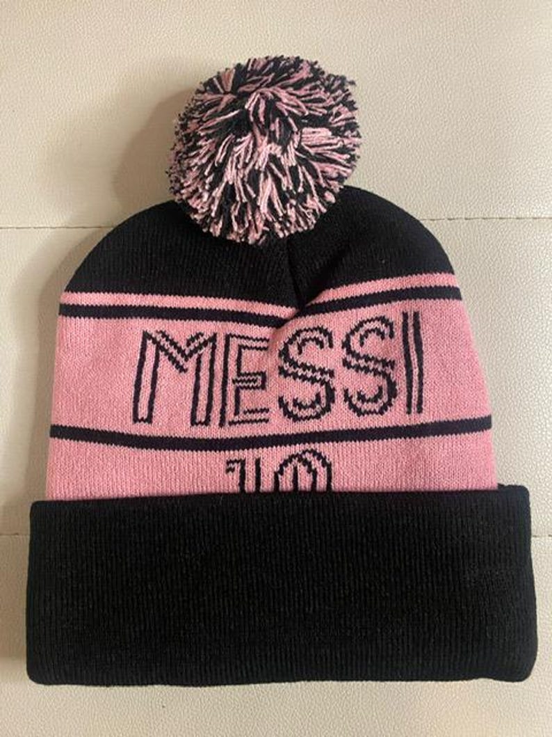 May include: Black and pink knit beanie with a pom pom on top. The beanie has the word "MESSI" and the number "10" embroidered on the front.