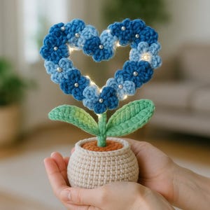 May include: A handcrafted crochet flower in a pot, featuring a heart-shaped arrangement of blue flowers with silver bead accents. The flower has a green stem and leaves, and the pot is beige. The flower is lit with small lights.