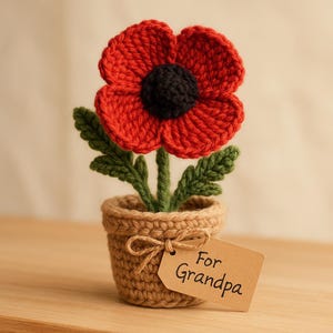 May include: A handmade crochet poppy flower in a small pot. The red flower has a black center and green leaves. The pot is tan with a bow and a tag that says "For Grandpa".