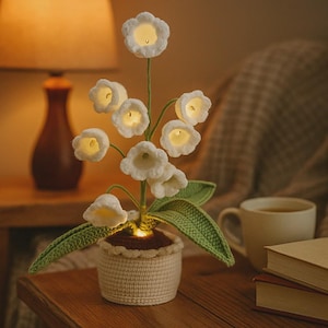 Puede incluir: Una lámpara de crochet de lirios del valle hecha a mano. Las flores blancas emiten un brillo suave y cálido. Las hojas y los tallos verdes están hechos a ganchillo, y la lámpara está en una maceta tejida. La lámpara está sobre una mesa de madera.