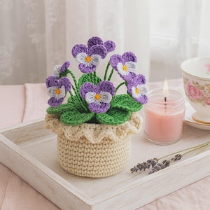Può includere: Una composizione floreale all'uncinetto con viole del pensiero viola e bianche con centri gialli e foglie verdi, in un vaso beige. I fiori fatti a mano sono su un vassoio bianco, accanto a una candela accesa e una tazza da tè.