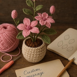 以下が含まれることがあります： 小さなベージュの鉢植えに手作りのかぎ針編みのフラワーアレンジメント。 ピンクの花には緑の葉があり、ピンクの糸玉、かぎ針編みフック、そして「Custom message here」と書かれたタグに囲まれています。