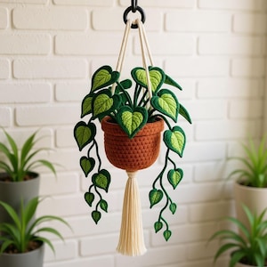 May include: A handcrafted, hanging crochet plant with a brown pot and green leaves. The plant is suspended by cream-colored rope and a black metal ring. A cream-colored tassel hangs below the pot. The leaves are green with lighter green accents.