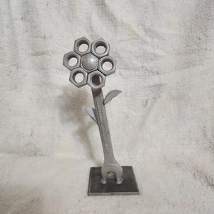 Puede incluir: Una escultura de flores de metal hecha a mano. Los pétalos de la flor están formados por seis tuercas hexagonales que rodean una esfera metálica central. El tallo es una llave inglesa y las hojas son pequeños recortes de metal. La escultura está montada sobre una base cuadrada de metal.