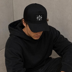 Könnte beinhalten: Schwarze Baseballkappe mit Mesh-Rückseite und einem quadratischen Aufnäher auf der Vorderseite. Der Aufnäher zeigt die Buchstaben "P A P A" in Weiß, in einem Kreuz angeordnet. Die Kappe wird von einer Person getragen, die einen schwarzen Kapuzenpullover trägt.