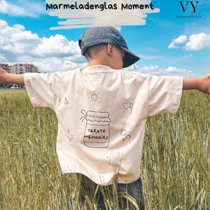 Könnte beinhalten: Cremefarbenes T-Shirt mit einem Glas-Motiv und der Aufschrift "CREATE MEMORIES" auf dem Rücken. Die Person, die das T-Shirt trägt, steht in einem Weizenfeld unter blauem Himmel. Der Text "Kreiere deinen Marmeladenglas Moment" ist darüber.