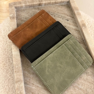 Könnte beinhalten: Drei minimalistische Kartenetuis in Braun, Schwarz und Salbeigrün. Jedes Portemonnaie hat mehrere Kartenfächer und einen genähten Rand. Die Geldbörsen sind auf einer neutralen Oberfläche angeordnet, um ihre Farben und Texturen hervorzuheben.