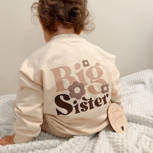 Könnte beinhalten: Ein cremefarbenes Sweatshirt mit den Worten "Big Sister" in braunen und rosa Blumenbuchstaben. Das Sweatshirt wird von einem Kind getragen. Ein Holzschild mit Text befindet sich neben dem Kind.