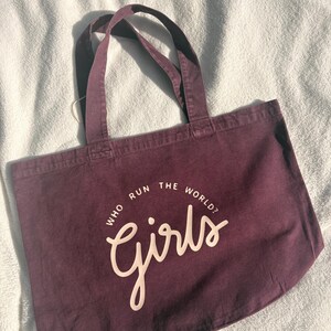 Op de afbeelding: Een donkerpaarse canvas draagtas met twee bijpassende hengsels. De voorzijde van de tas is voorzien van de tekst "WHO RUN THE WORLD?" in een klein, strak lettertype, met het woord "Girls" in een groot, lichtroze, cursief lettertype in het midden.