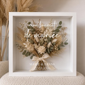 Op de afbeelding: Een vierkante witte schaduwdooslijst met een droogbloemenarrangement in beige, crème en groene tinten. Op het glas staat de witte tekst "mon bouquet de mariée" en "MARIE & FRANK 21.03.2026" boven het boeket dat is samengebonden met een crèmekleurig lint.