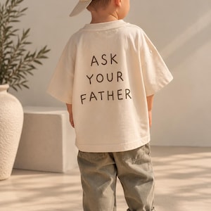 Peut inclure: Un t-shirt à manches courtes couleur crème avec le texte ASK YOUR FATHER imprimé en marron foncé au dos, associé à un pantalon vert olive, une casquette de baseball crème assortie et des baskets blanches, vu de dos.