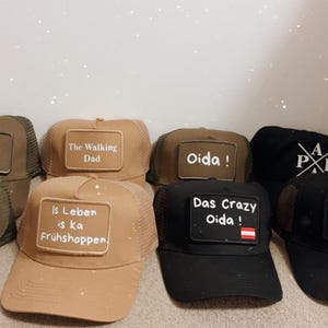 Könnte beinhalten: Eine Auswahl an Trucker-Caps in verschiedenen Farben, jeweils mit einem rechteckigen Aufnäher mit Text. Die Caps haben Mesh-Rücken und gebogene Schirme. Auf den Aufnähern stehen Sätze wie "The Walking Dad" und "Das Crazy Oida!"