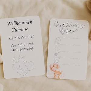Op de afbeelding: Twee witte kaarten met Duitse tekst en illustraties, waarschijnlijk voor een baby-aankondiging. Eén kaart zegt "Willkommen Zuhause" en de andere zegt "Unser Wunder ist geboren." Een knuffelkonijn en een teddybeer staan op de achtergrond.