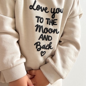 Könnte beinhalten: Beiges Sweatshirt mit dem Schriftzug "Love you to the moon and back" in erhabenen schwarzen Buchstaben. Das Sweatshirt hat einen Rundhalsausschnitt und lange Ärmel. Unter dem Text befindet sich ein kleines schwarzes Herz. Das Kind trägt eine passende beige Ripphose.