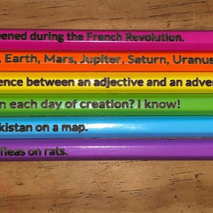 May include: Six colorful pencils with text, each in a different color: pink, orange, yellow, green, blue, and purple. The pencils have green erasers and silver metal bands. Each pencil has a different phrase printed on it, such as "Ask me what happened during the French Revolution."