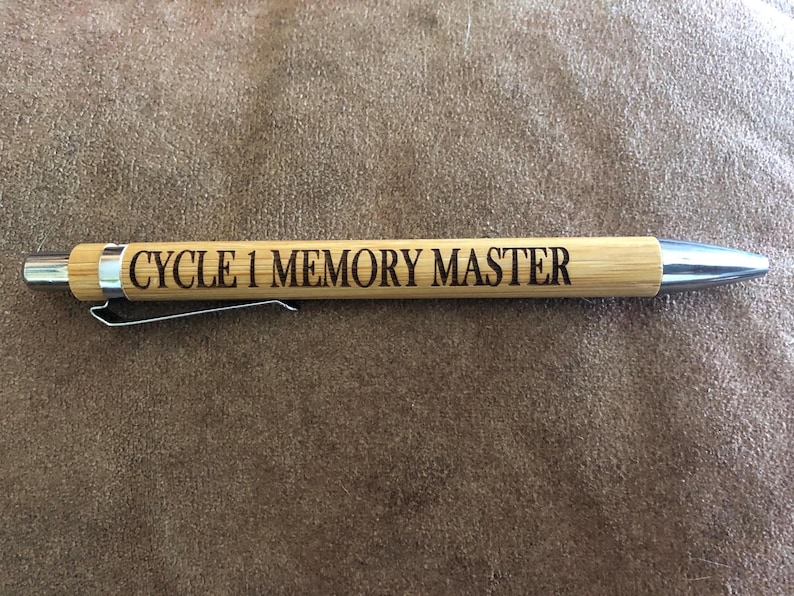 Pu&ograve; includere: Una penna in legno marrone con una clip argentata e una punta argentata. La penna &egrave; incisa con il testo "CYCLE 1 MEMORY MASTER".