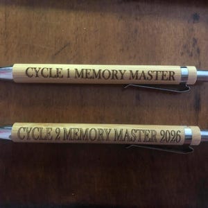 May include: Two bamboo pens with silver accents. The pens have the text "CYCLE 1 MEMORY MASTER" and "CYCLE 2 MEMORY MASTER 2026" engraved on them. The pens are lying on a brown wooden surface.