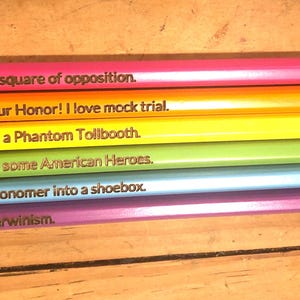 May include: Six colorful pencils with humorous sayings printed on them. The sayings are: "I can draw the square of opposition", "Objection, Your Honor! I love mock trial", "I took a ride in a Phantom Tollbooth", "Ask me about some American Heroes", "I stuck an astronomer into a shoebox", and "I defeated Darwinism."