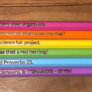 May include: Six colorful pencils with humorous phrases printed on them. The pencils are arranged in a row, each a different color: pink, orange, yellow, green, blue, and purple. The text on the pencils includes phrases like "I can show you where your organs are."