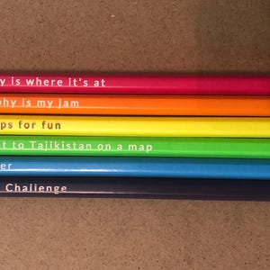 May include: A collection of six pencils in a rainbow of colors, each with a different geography-themed phrase printed on the side. The pencils have pink, orange, yellow, green, blue, and purple barrels, with matching erasers.