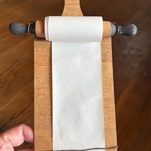 May include: A wooden wall-mounted notepad holder featuring a light brown paddle-shaped board with a hole at the top. A miniature rolling pin with dark gray handles holds a roll of white paper that extends down the length of the board.