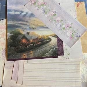 May include: A collection of decorative paper sheets featuring various designs, including a landscape scene with a cottage, a floral border with purple and pink hydrangea blossoms, striped patterns, and solid textured backgrounds in muted tones.