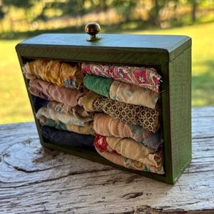 May include: A rectangular green wooden drawer with a small brass knob, containing multiple folded strips of fabric in various colors and patterns, including yellow, pink, black, and floral prints, stacked neatly inside the open frame.