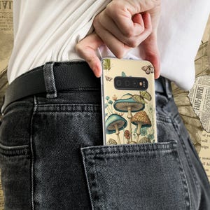 Peut inclure: Une coque de t&eacute;l&eacute;phone avec un fond beige et une illustration color&eacute;e de champignons et de papillons. La coque est dans une poche arri&egrave;re d'un jean fonc&eacute;.