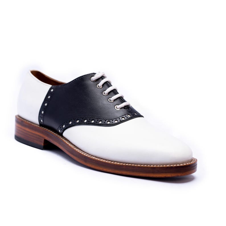 May include: A white and black saddle shoe with a brown sole. The shoe features a black leather saddle and white laces. The shoe has a classic design and is likely made of leather.