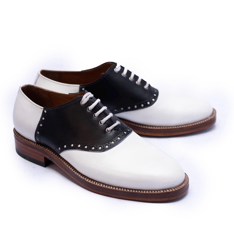 May include: A pair of classic saddle shoes in black and white leather. The shoes feature white laces, a brown sole, and decorative stitching. The black leather section is on the top of the shoe, with white leather on the toe and heel.