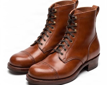 May include: A pair of tan leather lace-up boots featuring a cap toe with decorative brogue perforations. The boots have dark brown laces, brass-toned eyelets, a stacked heel, and a pull tab at the back of the ankle collar for easy access.