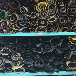 May include: Shelves filled with rolled leather hides in various colors, including green, purple, and black. The rolls are neatly organized on blue shelves, creating a visually appealing display of materials.
