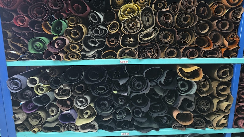 May include: Shelves filled with rolled leather hides in various colors, including green, purple, and black. The rolls are neatly organized on blue shelves, creating a visually appealing display of materials.