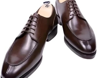 Könnte beinhalten: Ein Paar braune Lederschuhe mit klassischem Design. Die Schuhe haben eine polierte Oberfläche, eine runde Spitze und einen Schnürverschluss. Die Sohlen sind schwarz und die Schuhe werden auf hölzernen Schuhspannern präsentiert.